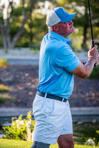 Man in blue shirt and white shorts playing golf on a grassy course with trees in the background