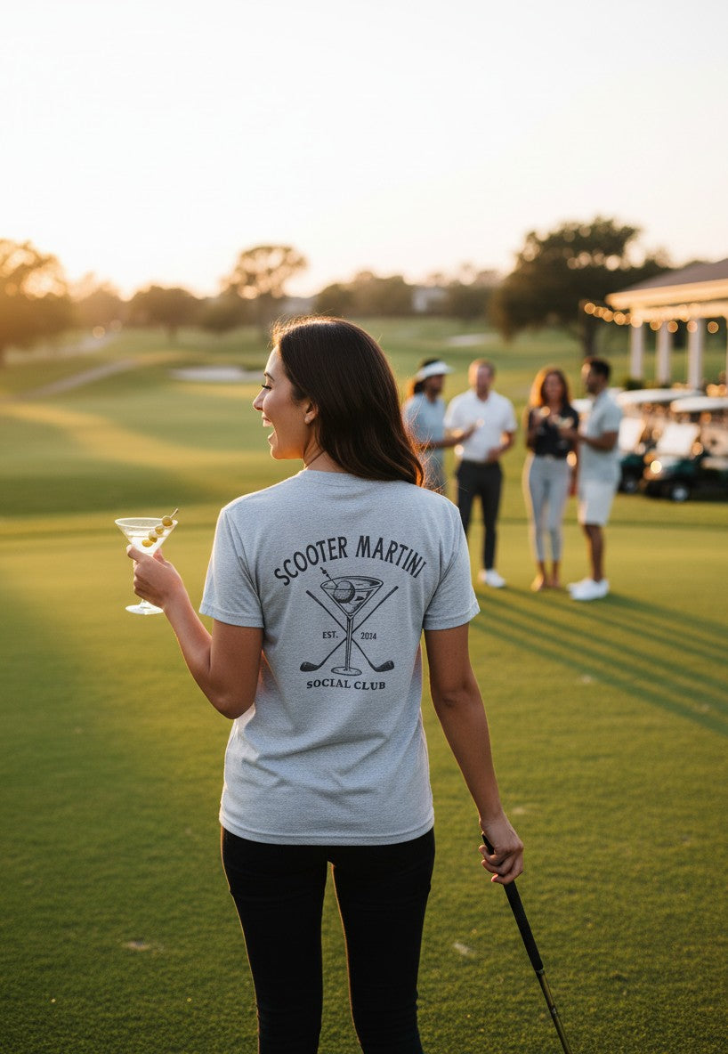Woman wearing a heather white Scooter Martini tri-blend t-shirt with a golf-themed martini logo on a golf course.